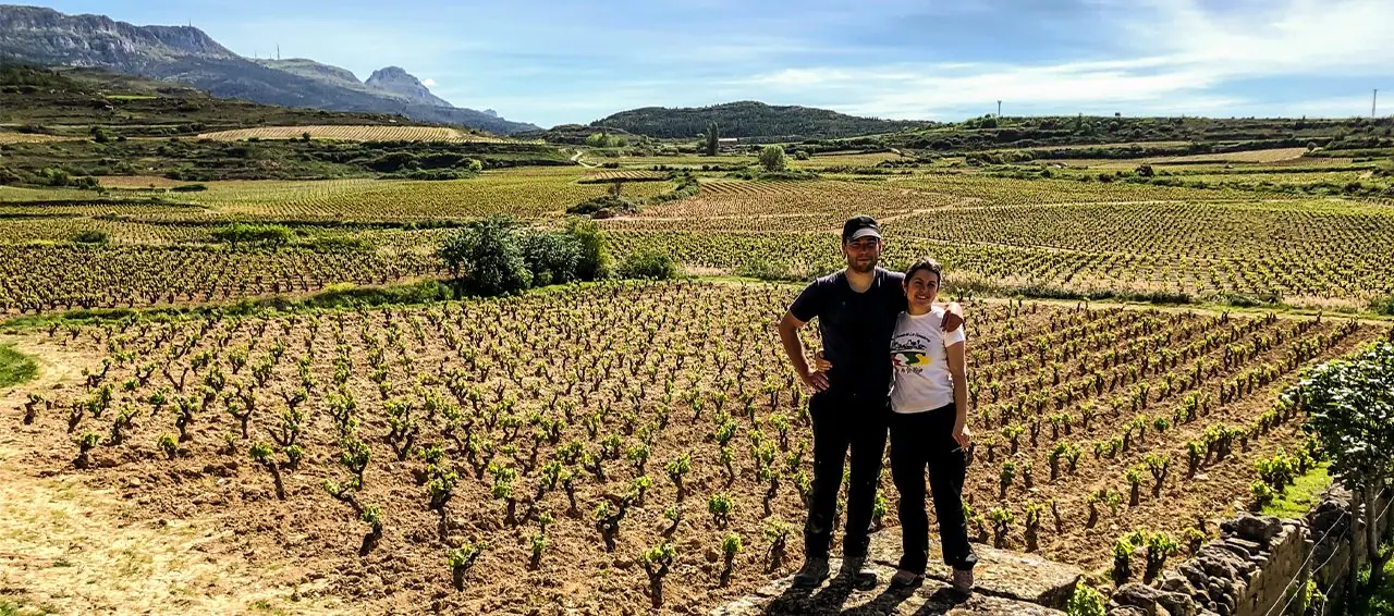 Un hombre y una mujer de pie sobre un muro de piedra ante un extenso viñedo de cepas en vaso recién brotadas, con una sierra rocosa al fondo.